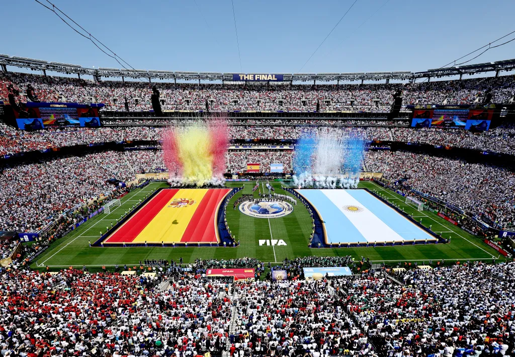 General view of a stadium filled with fans, showing large flags of Spain and Argentina on the field with pyrotechnics.