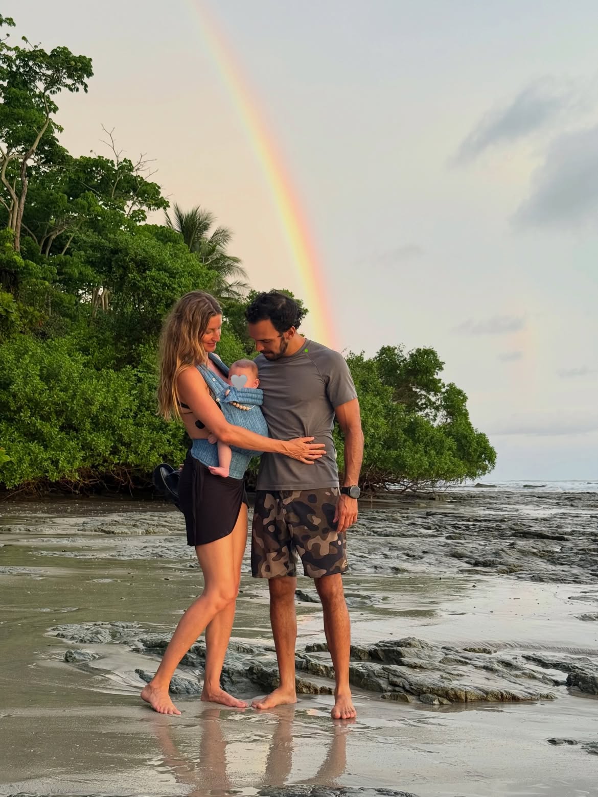 Gisele Bündchen, Joaquim Valente, and a baby standing on a rocky beach under a rainbow.