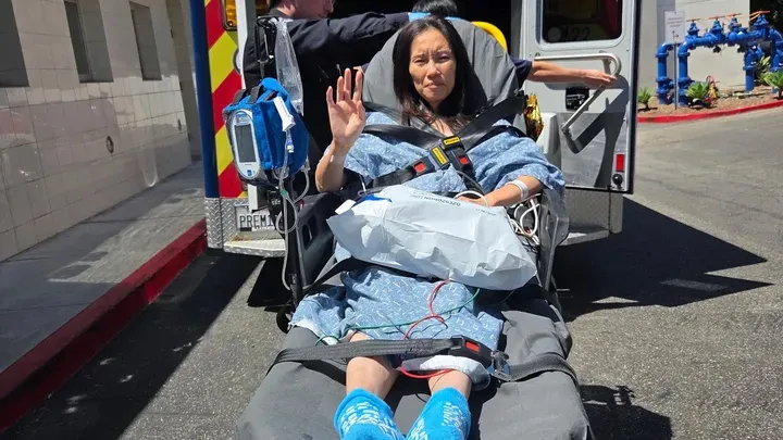 A woman in a hospital gown and blue slippers is strapped to a stretcher, waving, as she's loaded into an ambulance.