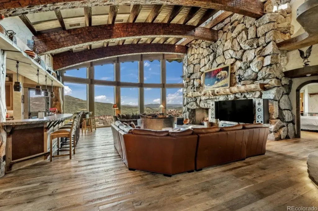 A great room in a luxury mansion with a stone fireplace, brown sectional sofa, and large windows with mountain views.