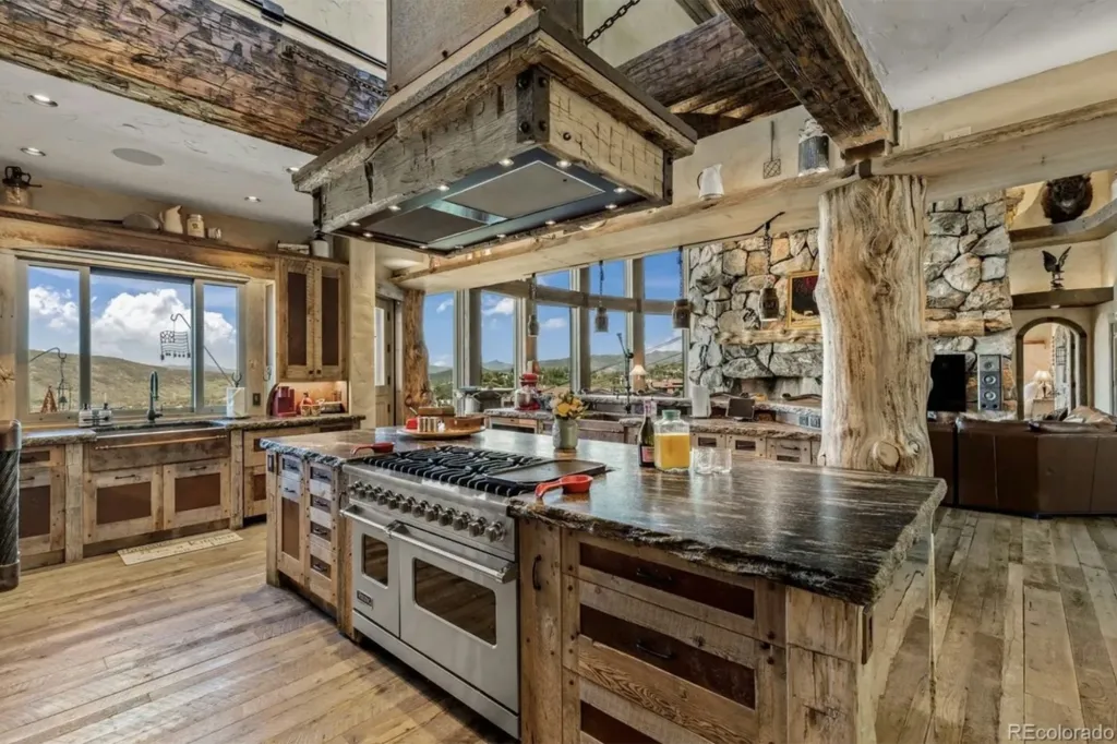 A rustic kitchen with a large island, wooden beams, a stone fireplace, and mountain views from large windows.