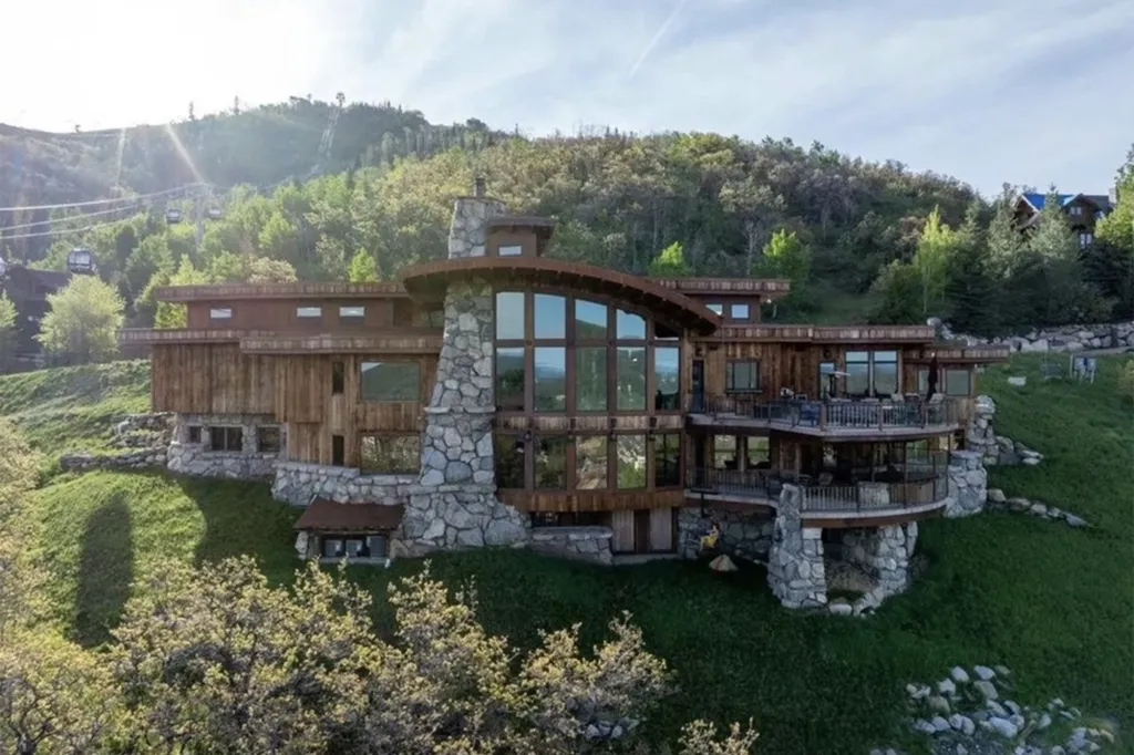 A large stone and wood mansion with multiple balconies and large windows on a green hillside with gondolas visible in the background.