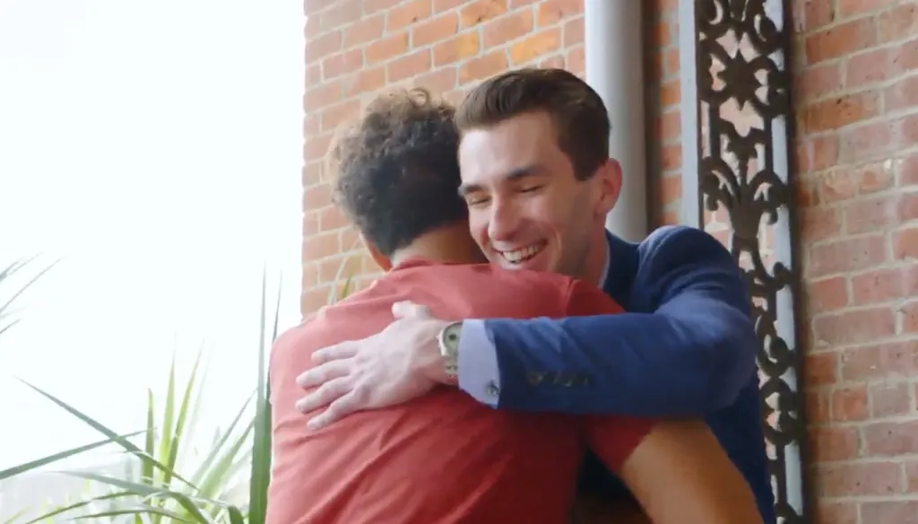 A smiling man in a blue suit hugs a man in a red shirt.