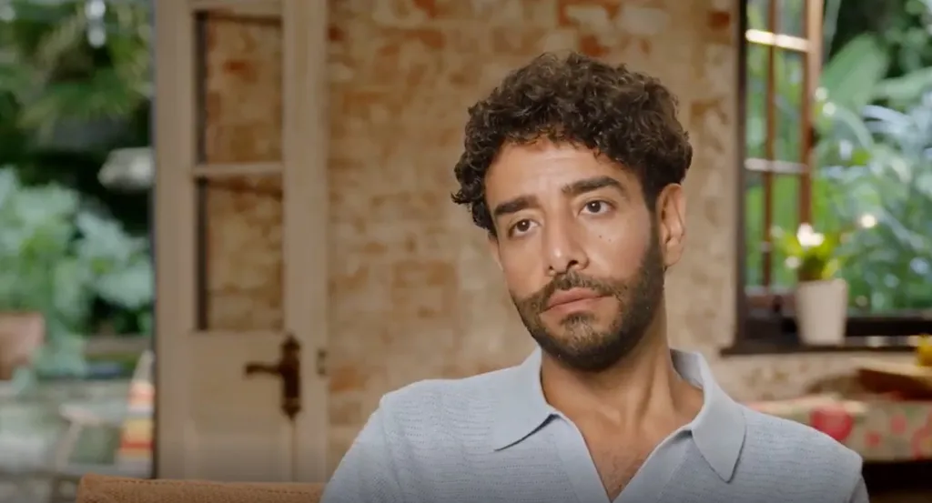 A man with curly hair and a beard, wearing a light blue polo shirt, looks to the right.
