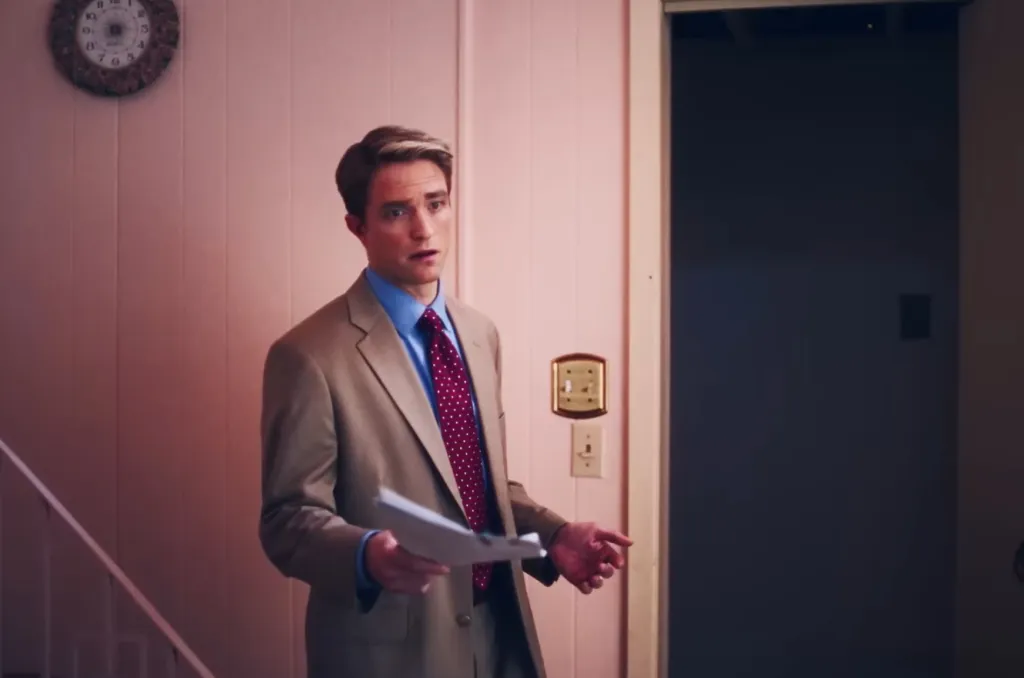 Robert Pattinson as Chris Hansen, wearing a tan suit, blue shirt, and red patterned tie, looking surprised.
