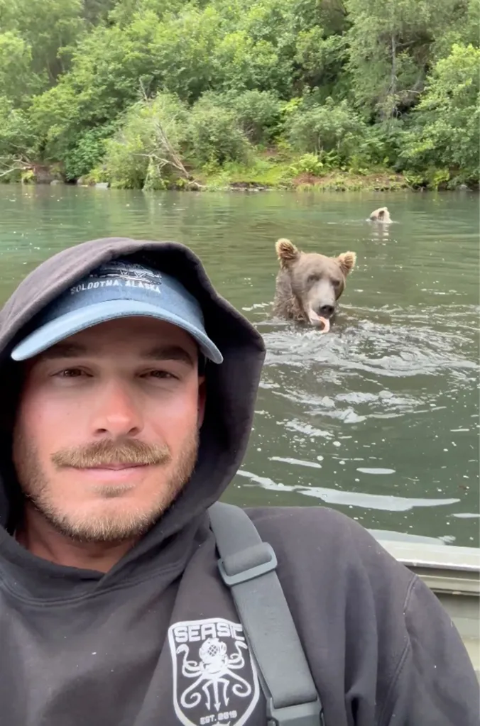 A man in a boat takes a selfie with a grizzly bear in the water with a fish in its mouth.