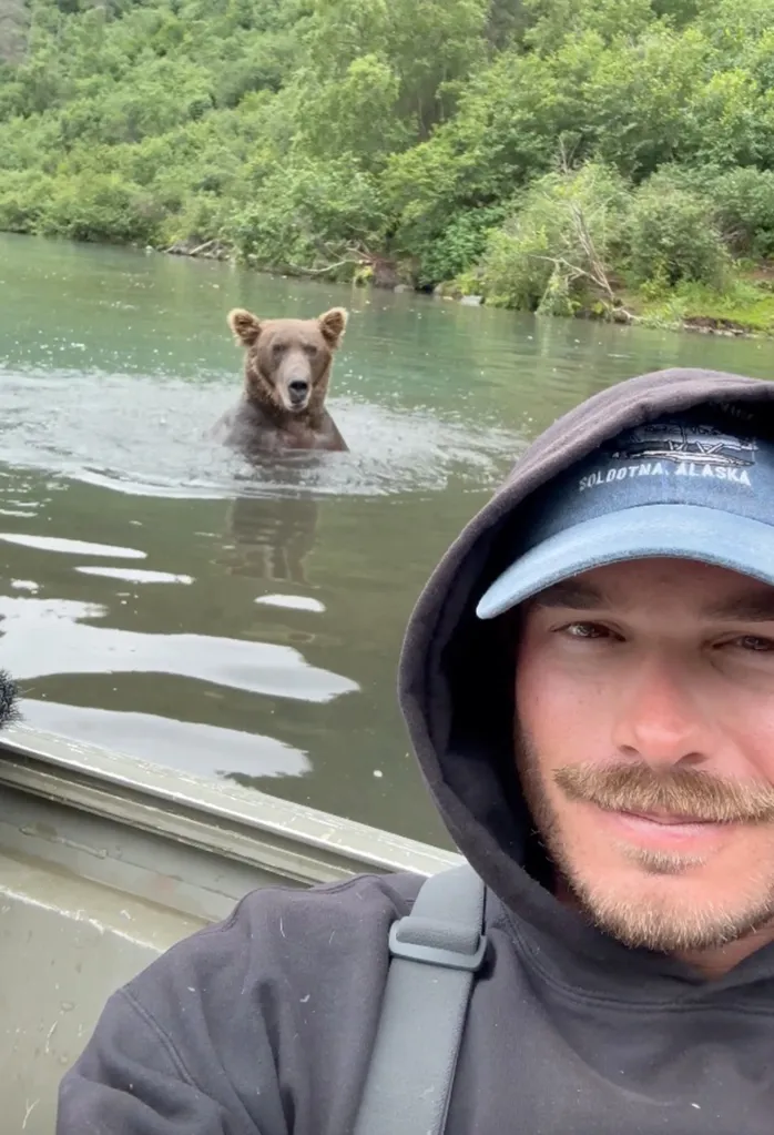 A man in a boat takes a selfie with a grizzly bear in the water behind him.