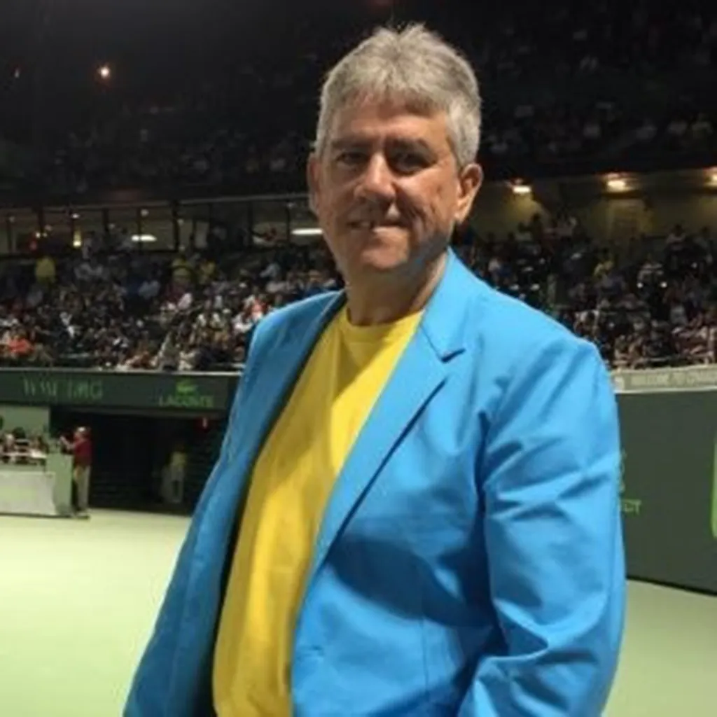 Gustavo E. Gordillo, father of Gustavo Gordillo, in a blue blazer and yellow shirt at a sports stadium.