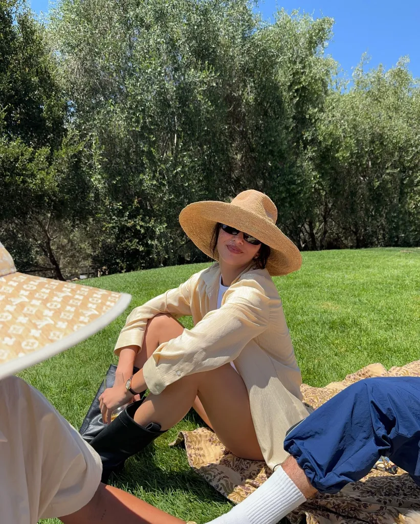 Kendall Jenner sitting on a patterned blanket in the grass, wearing a straw sun hat, sunglasses, and a light-colored top, with trees in the background.