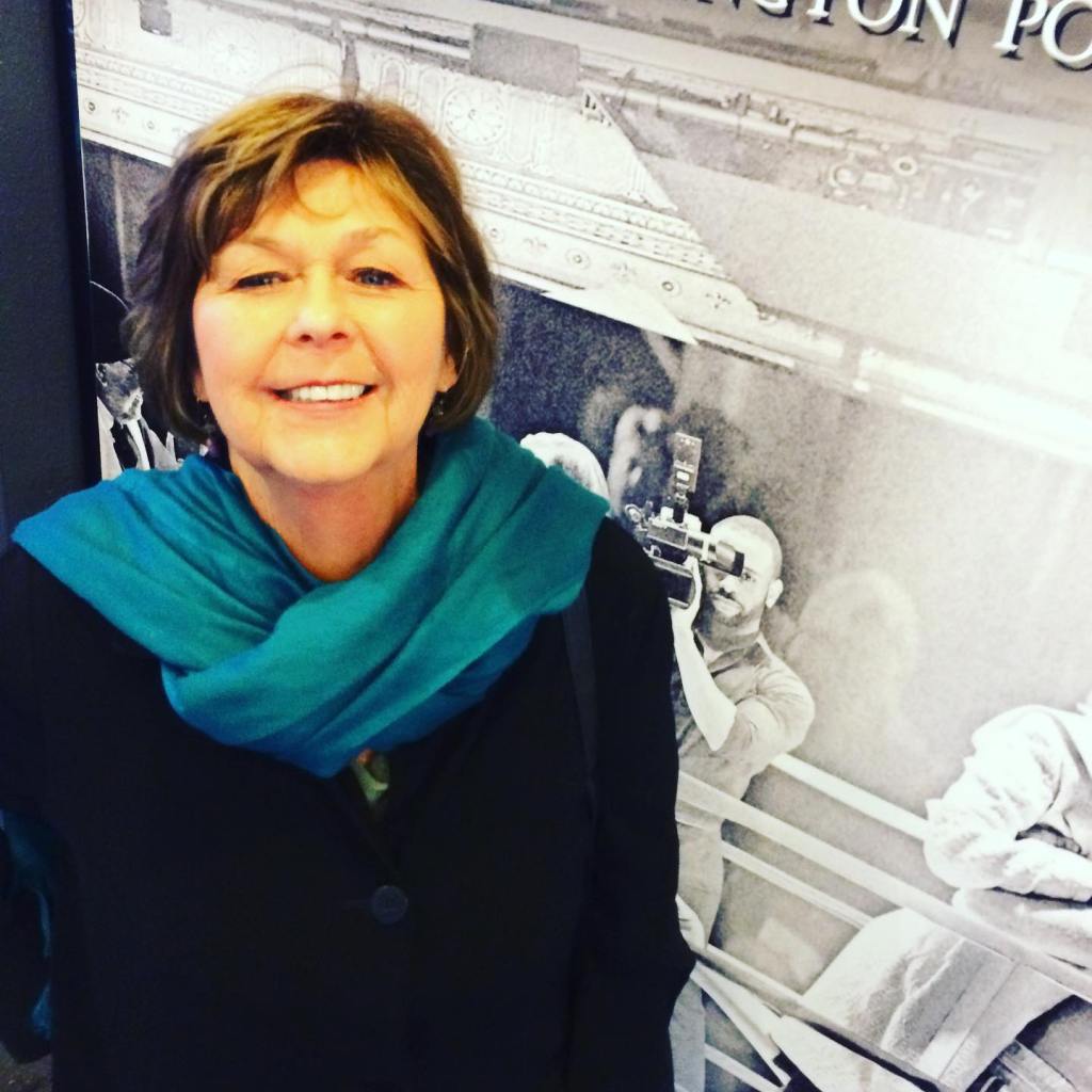A smiling Nancy Guthrie wearing a blue scarf in front of a black and white photo of a cameraman.