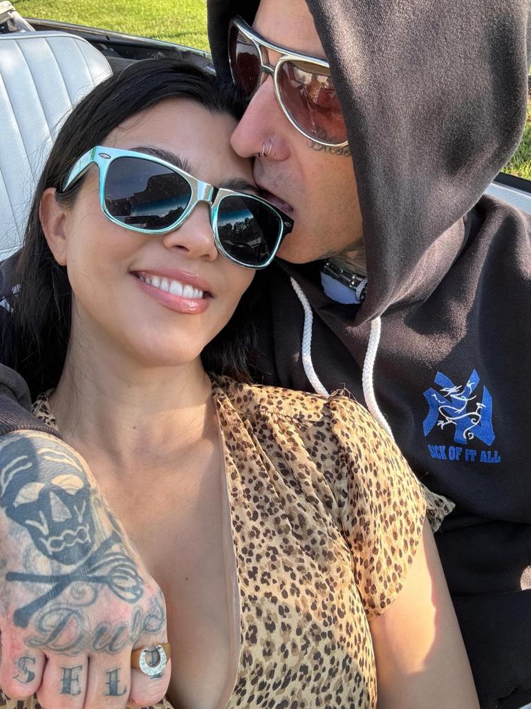 Kourtney Kardashian smiling in turquoise sunglasses as Travis Barker, in a black hoodie and sunglasses, kisses her forehead.