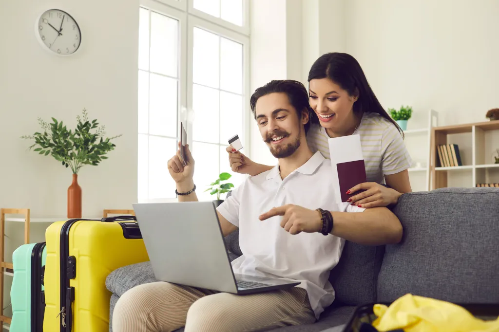 A happy couple uses a laptop to book a trip, holding a credit card and passport.
