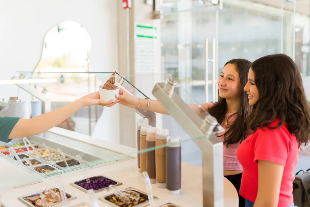 Two smiling teen girls receive ice cream from a store worker.