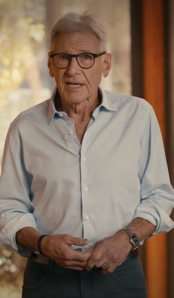 Harrison Ford in a light blue button-up shirt and glasses, speaking to the camera.