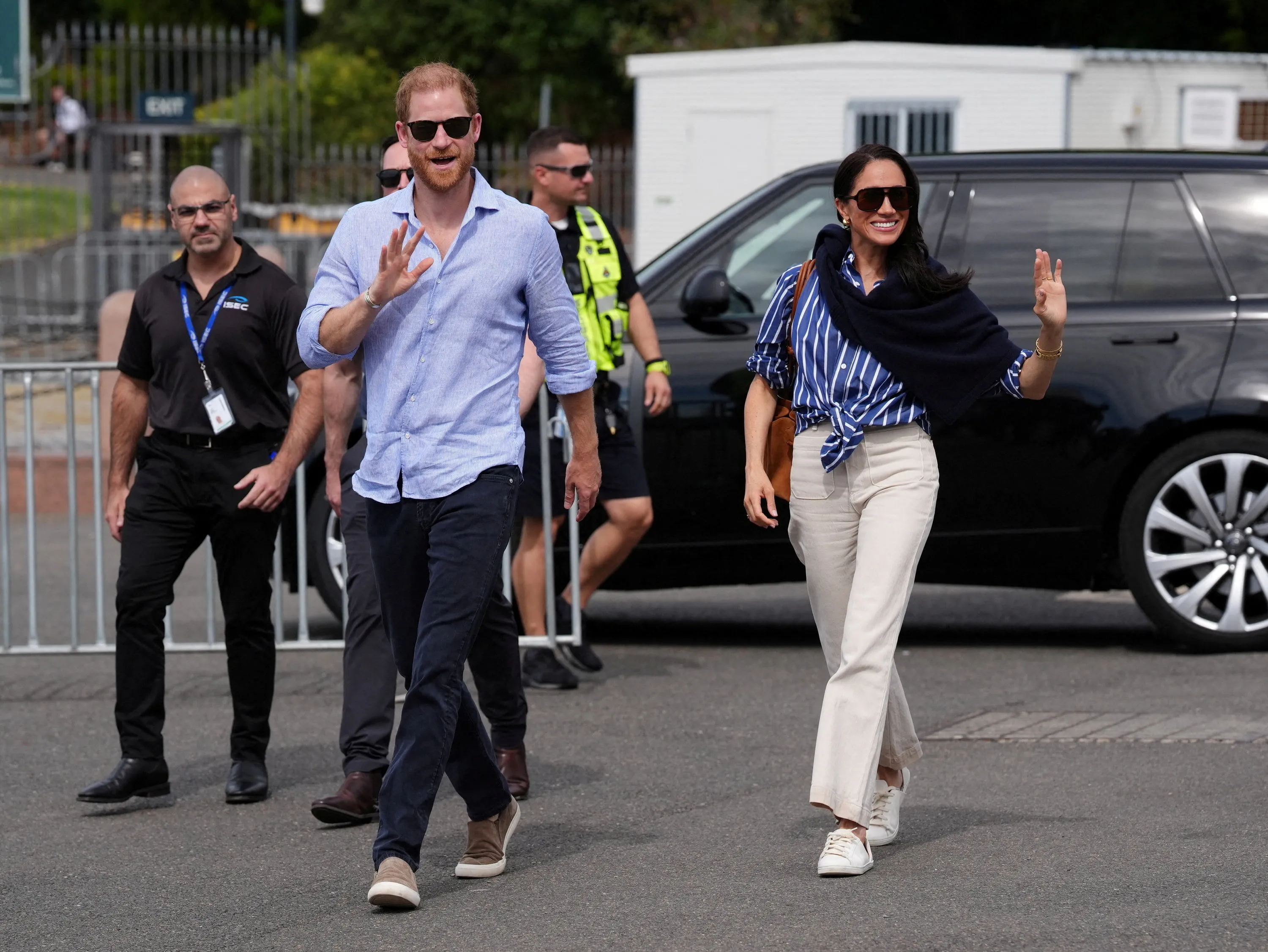 Prince Harry and Meghan Markle wave to the crowd as they walk past a car and security personnel.