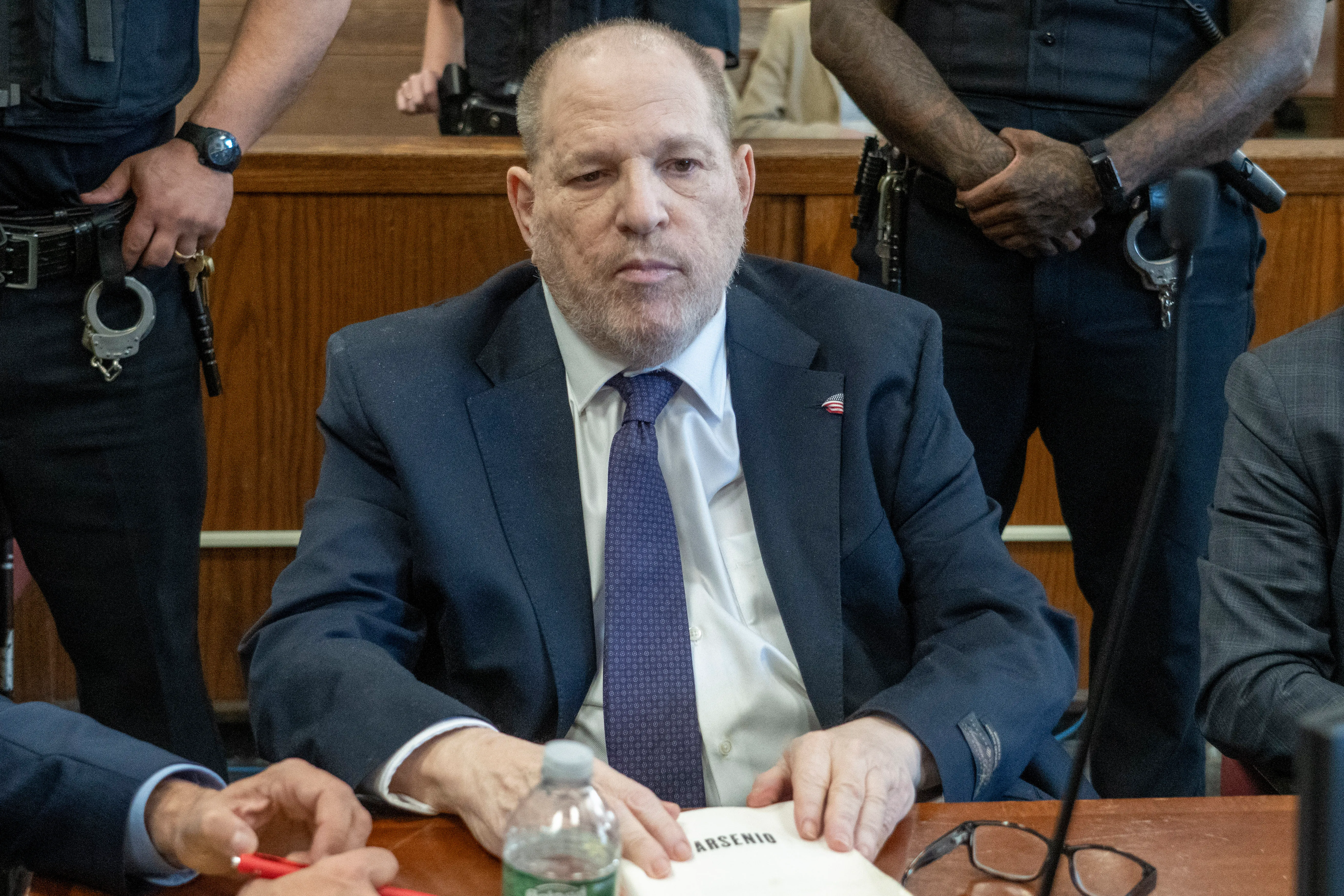 Harvey Weinstein at his trial, seated at a table with a police officer standing behind him.