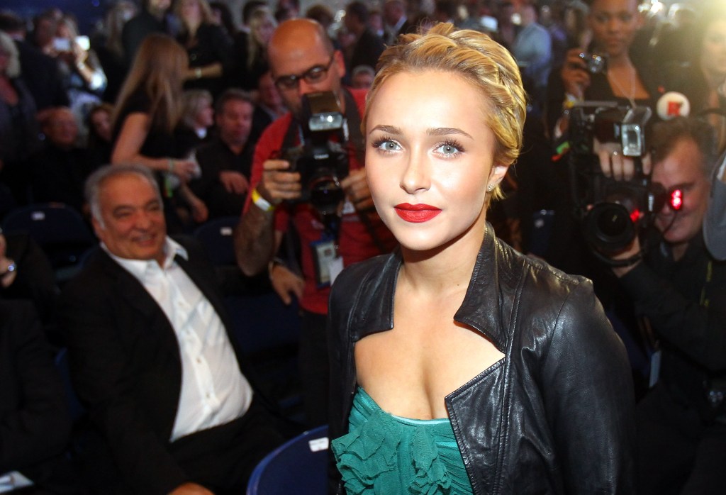 Hayden Panettiere wearing a black leather jacket over a green ruffled top, with a man smiling in the background and photographers in the back.