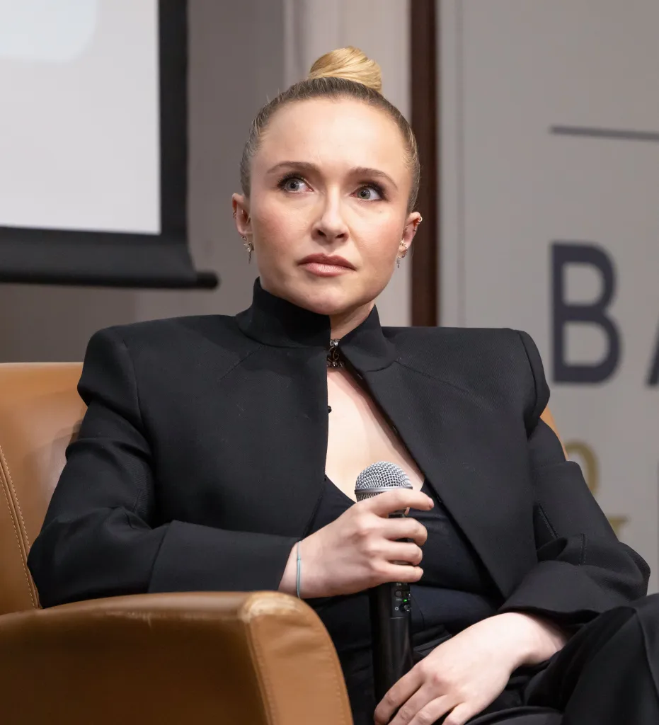 Hayden Panettiere in a black suit with hair in a bun holds a microphone while looking up and to the right.