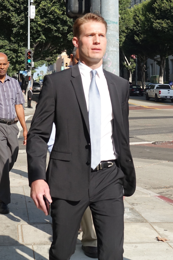 Brian Hickerson in a black suit with a light blue tie, walking on a sidewalk.