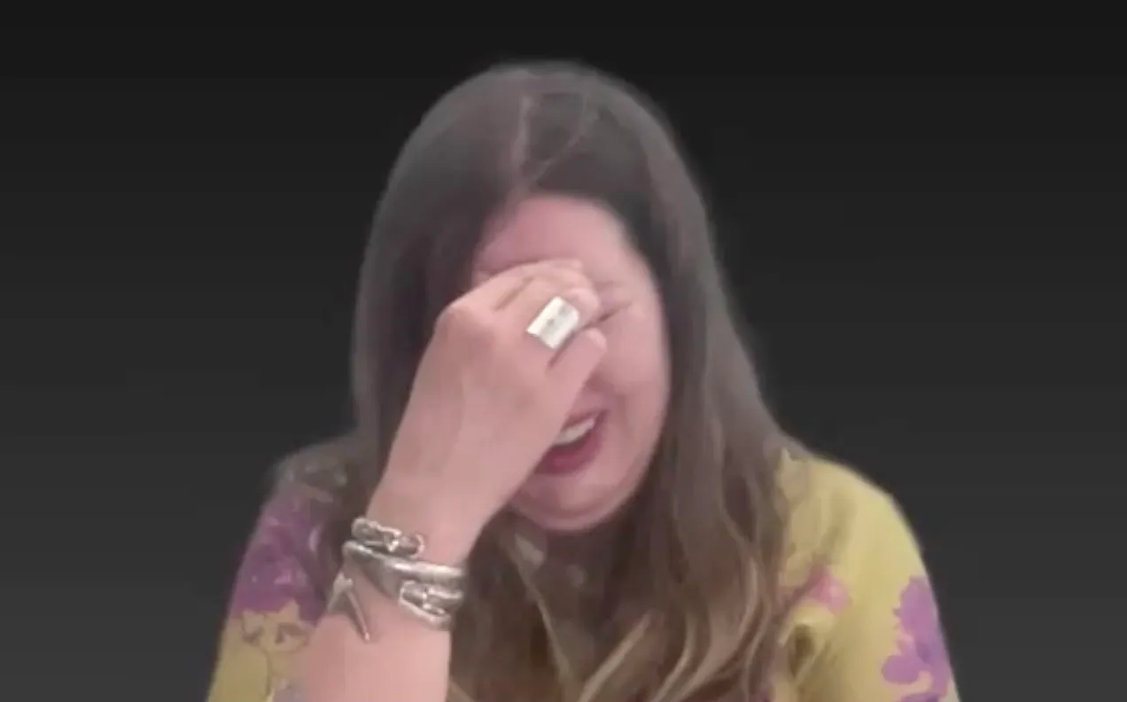 A woman with long brown hair, wearing a patterned top and bracelets, covering her face with one hand.