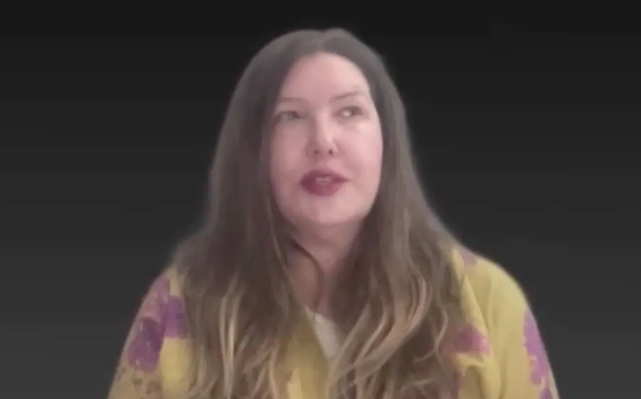 A woman with long wavy brown hair and red lipstick on a dark background.