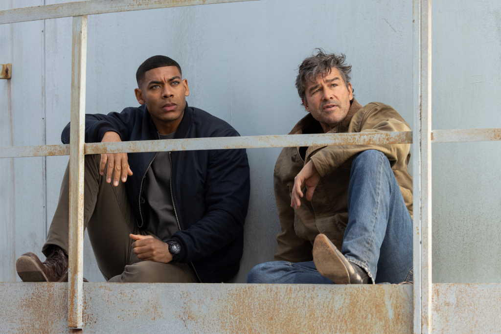 Two men, one Black and one white, sitting behind a rusty metal railing.