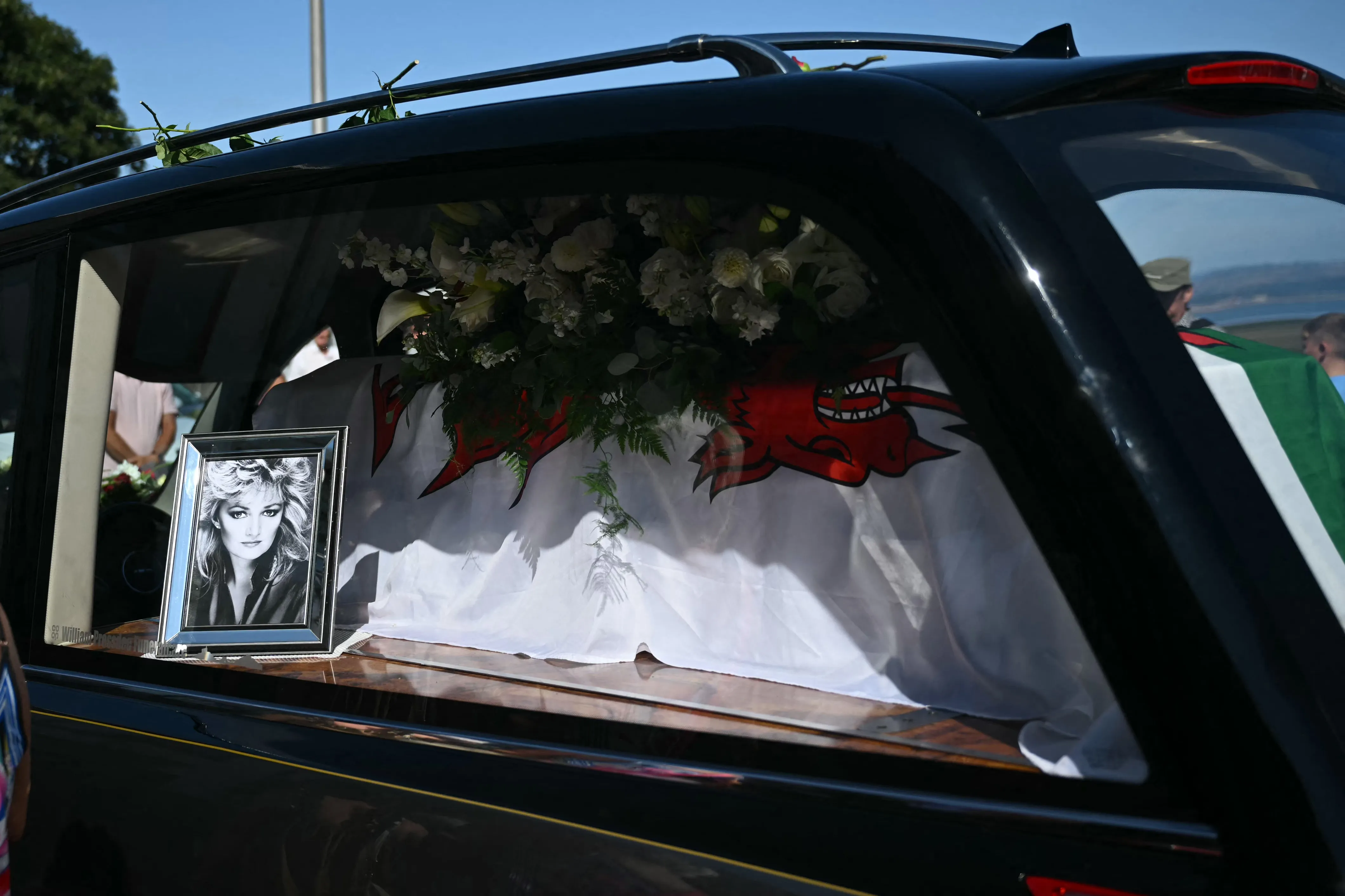 A hearse carrying the coffin of Bonnie Tyler, draped with a Welsh flag and flowers, with a framed photo of the singer.