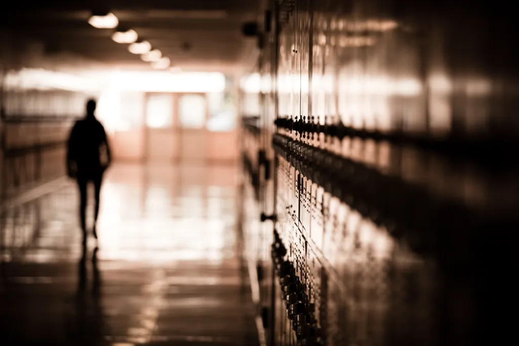 A high school student walks down a dark hallway, silhouetted by daylight.
