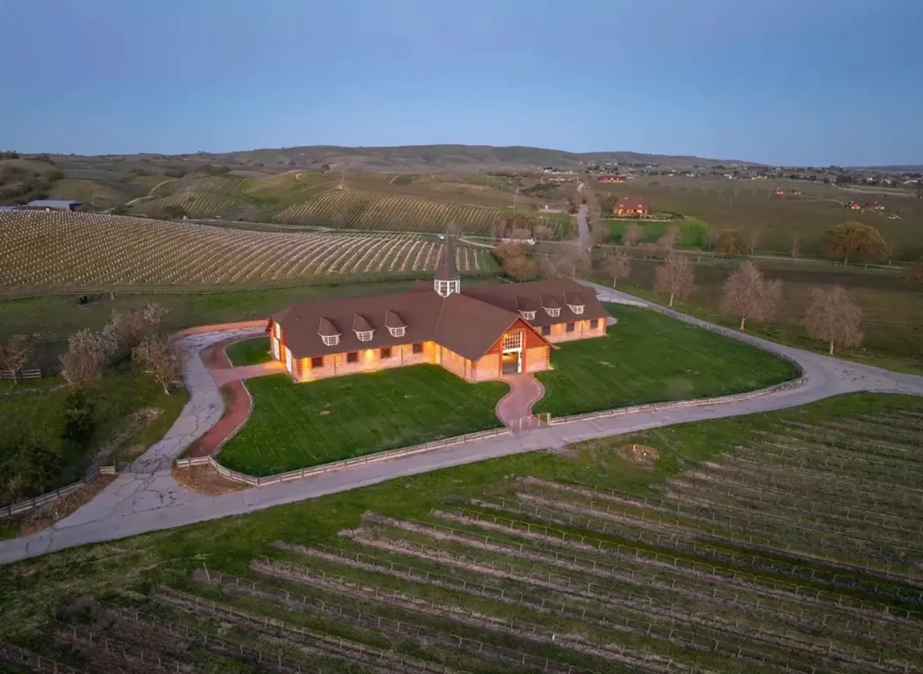 Aerial view of Windfall Farms, a 700-acre estate with vineyards, hills, and a large building with a brown roof and lit windows in the center.