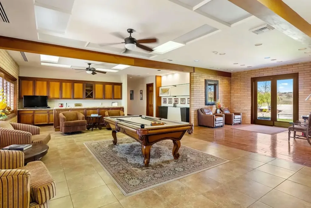A large room with a pool table on an area rug, various armchairs, and a built-in wooden cabinet with a TV.