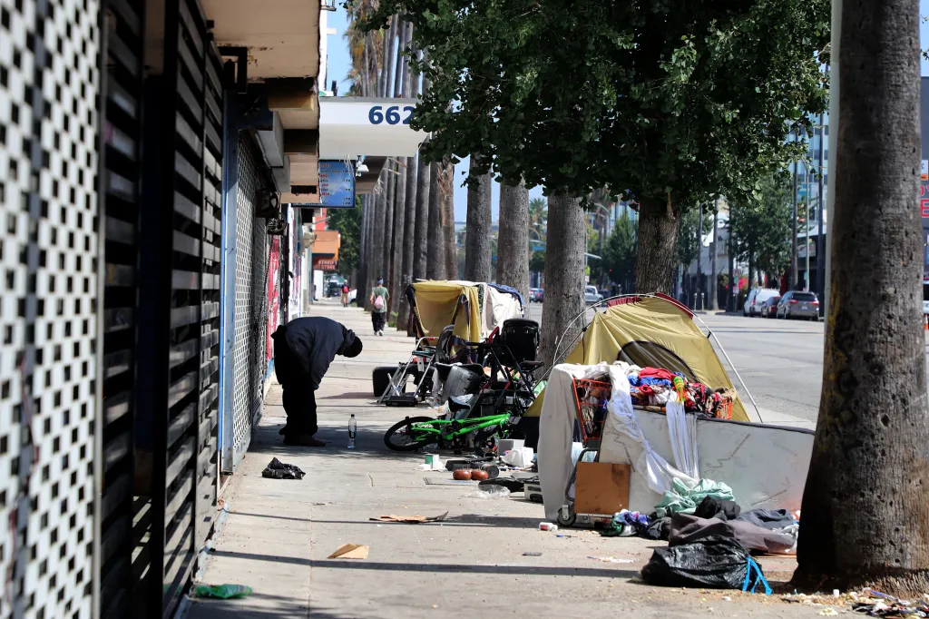 A person bending over near closed storefronts and homeless encampments on Van Nuys Blvd.