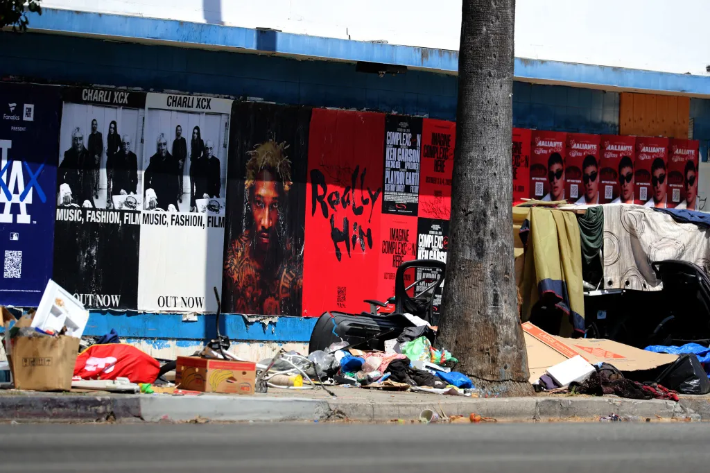 Overturned office chair and trash next to a tree trunk on a street curb in front of a wall covered in posters.