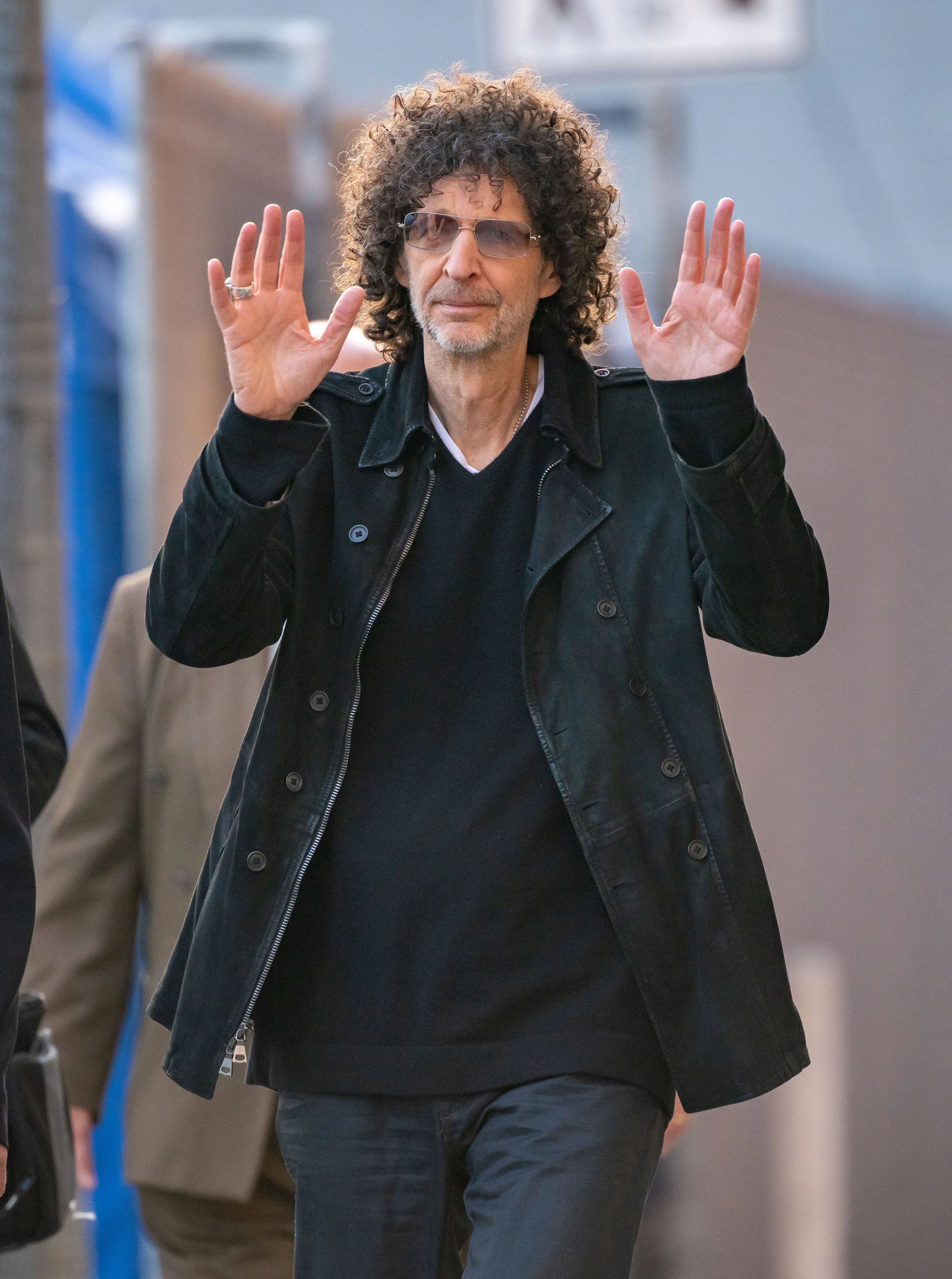 Howard Stern raises both hands while wearing a dark jacket and sunglasses.