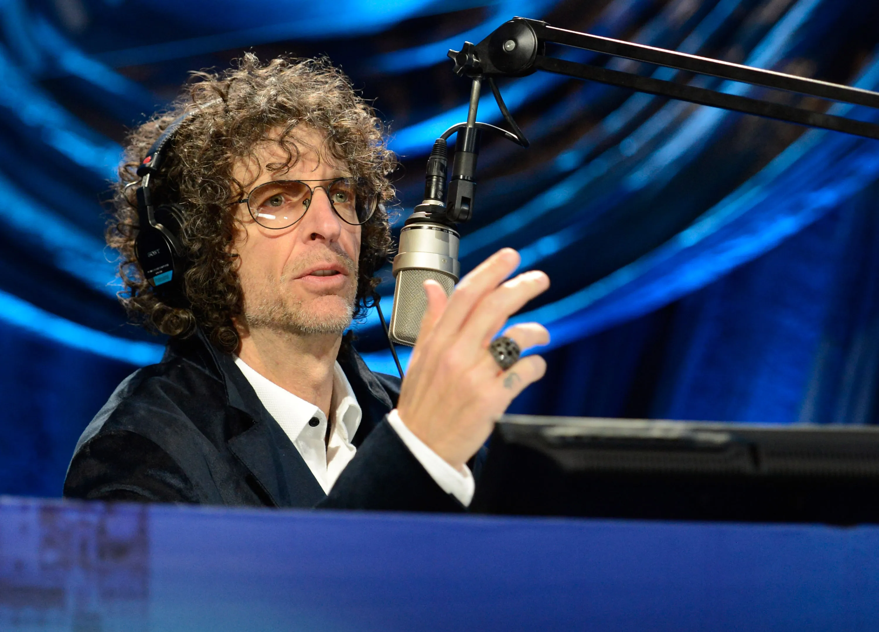 Howard Stern wearing headphones and glasses, speaking into a microphone.