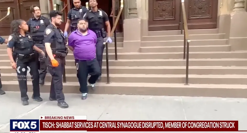 Four police officers escort a man in a purple shirt, who is in handcuffs, down the steps of a building.