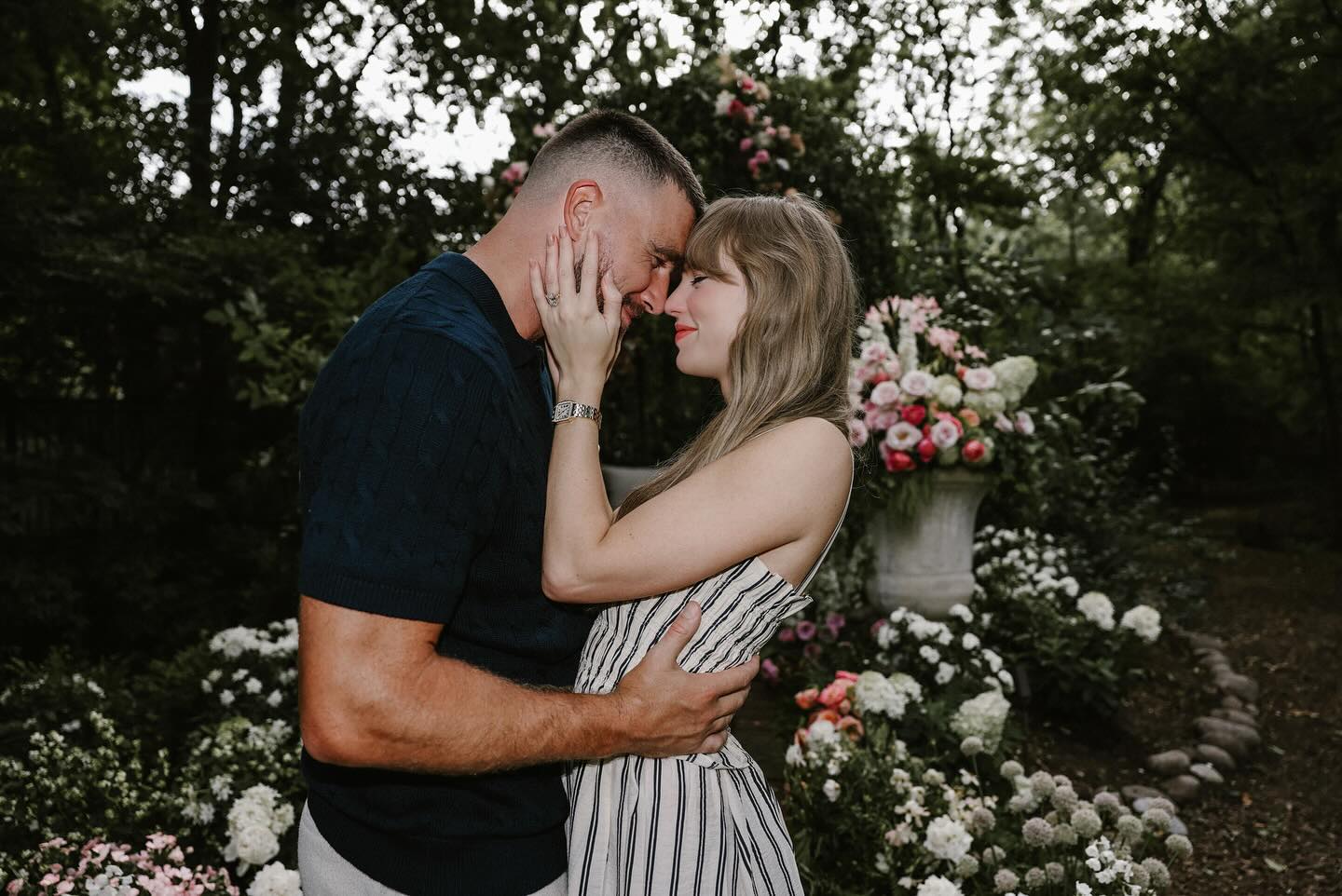 Taylor Swift and Travis Kelce embracing with foreheads touching, surrounded by flowers.