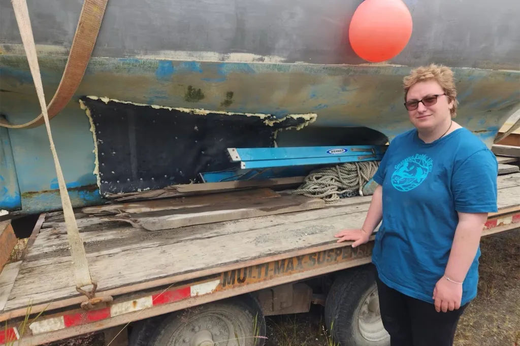 A person standing next to a large boat with hull damage on a flatbed trailer.
