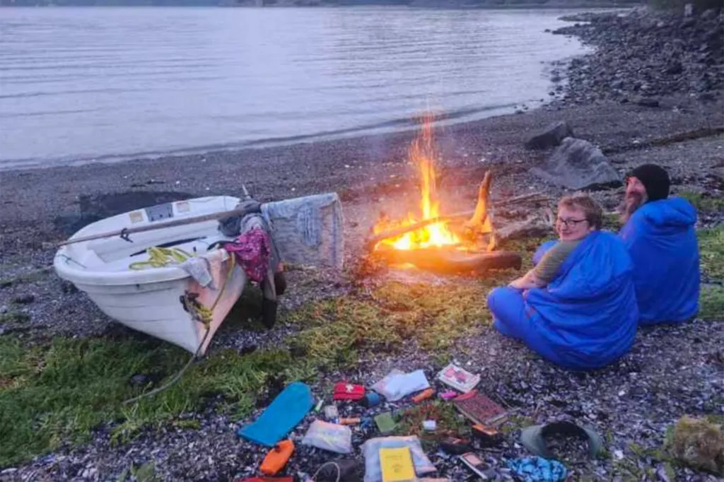 Two people in blue sleeping bags sit by a campfire next to a rowboat on a rocky beach.