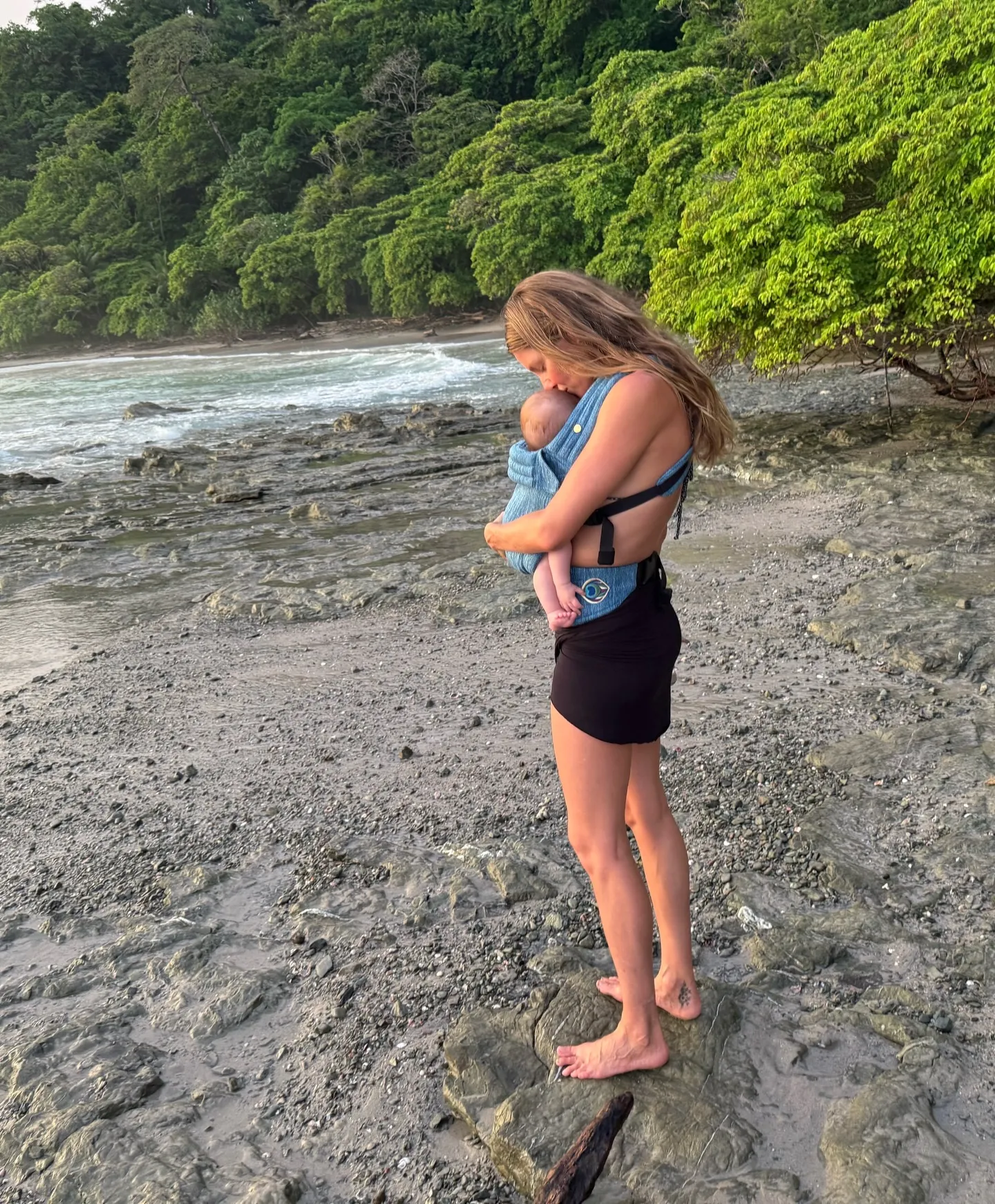 Gisele Bündchen holding her baby in a carrier on a rocky beach.