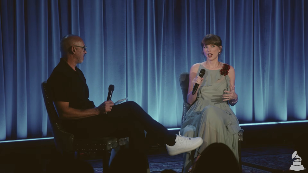 Taylor Swift speaking into a microphone while seated next to a man with a microphone during an interview.