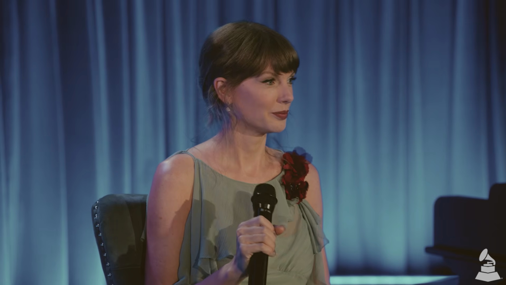 Taylor Swift, wearing a light green dress with a red flower on her shoulder, holds a microphone and looks to the right.
