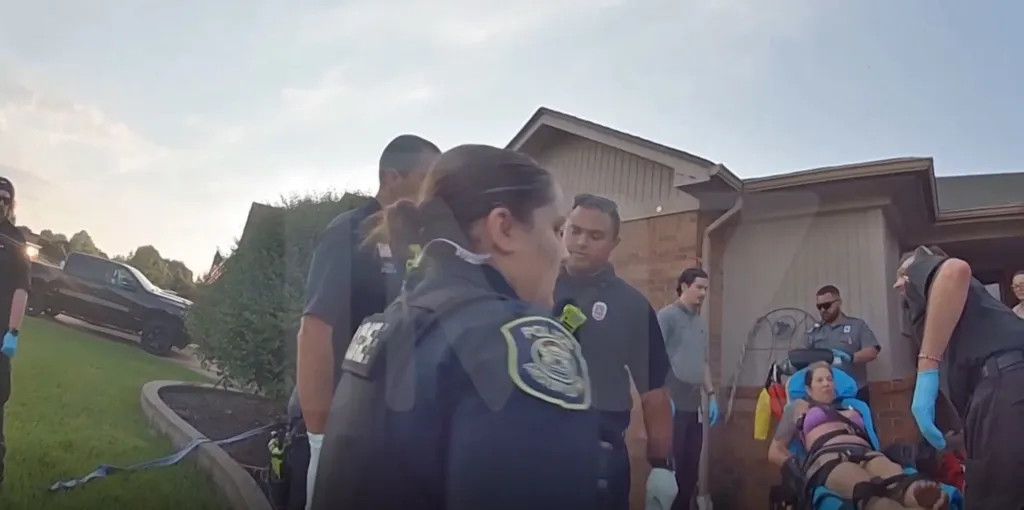 Police and paramedics assisting a woman on a stretcher outside a house.
