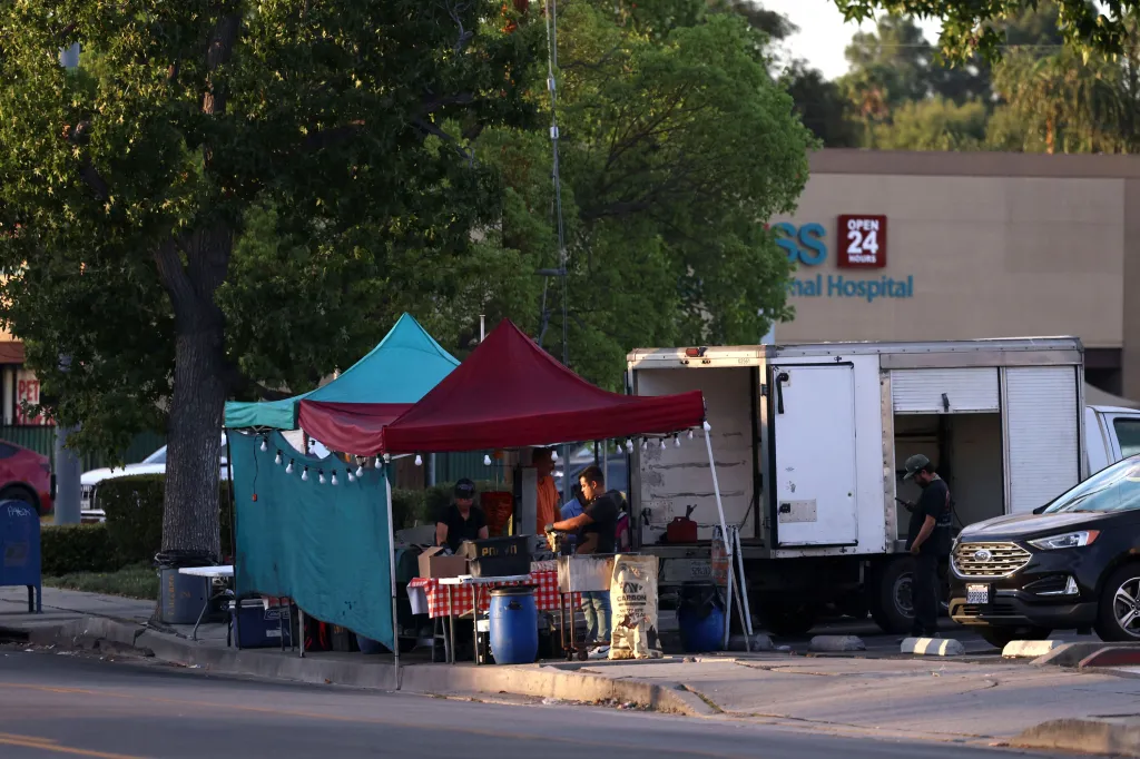 Illegal street vendors operate under canopies next to a street, with a hospital in the background.