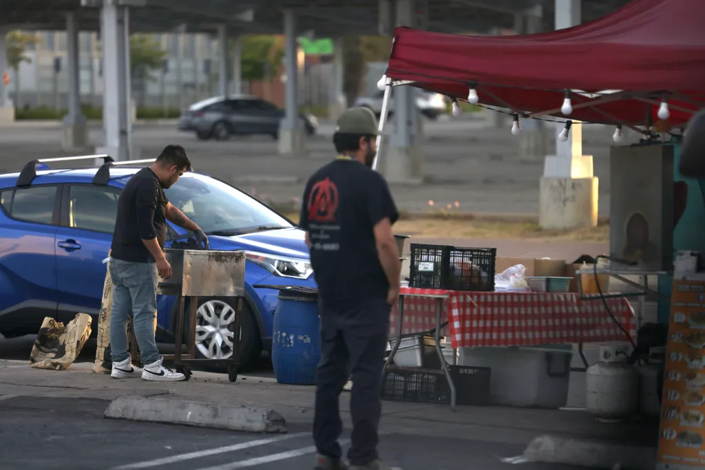 Street vendors selling goods from under a red tent and next to a blue car.