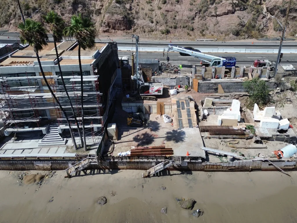 Aerial view of a Malibu construction site on a beach, rebuilding a home destroyed by wildfires, with palm trees, scaffolding, heavy machinery, and construction materials visible.