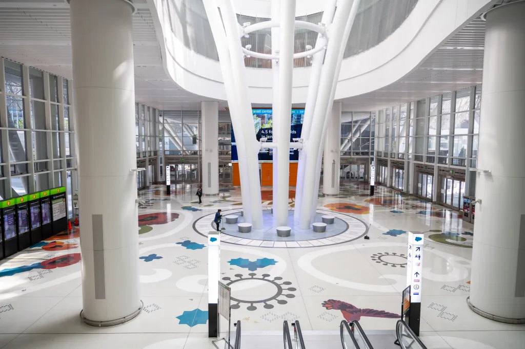 Interior of the Salesforce Transit Center grand hall with sculptural white columns, a colorful patterned floor, and sparse commuters.