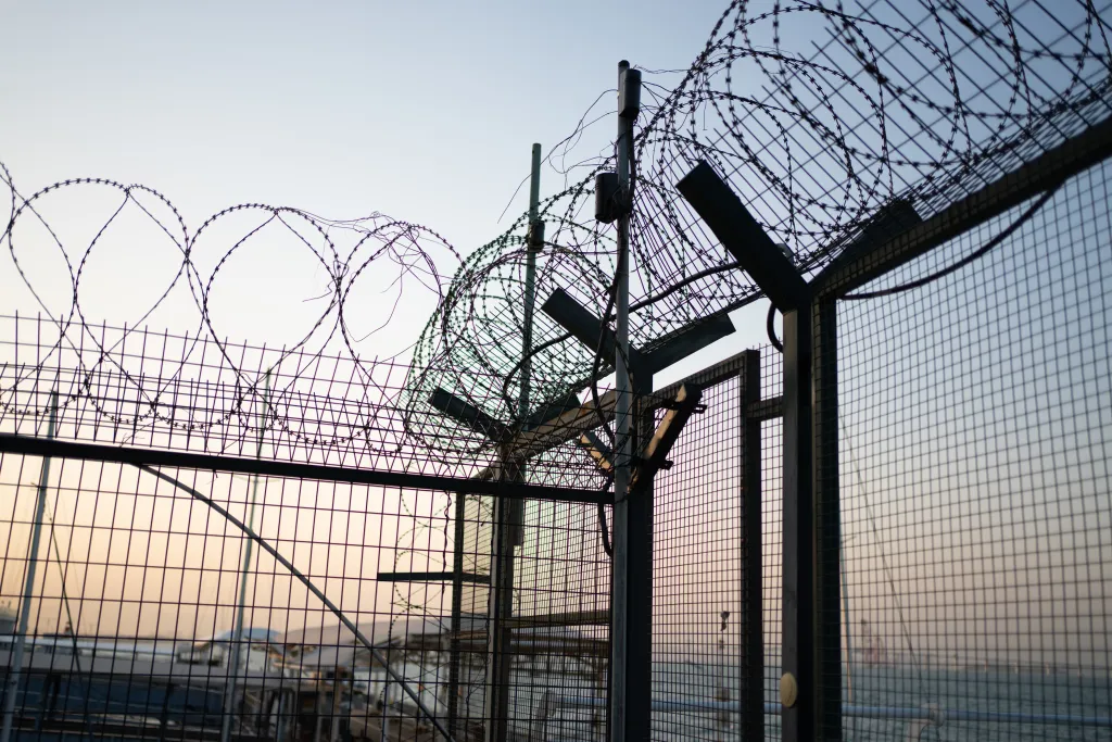 Iron mesh isolation area with razor wire on top, overlooking a hazy sunset.