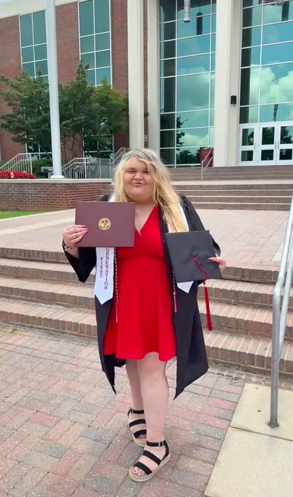 A woman in a red dress, graduation gown, and sandals holds a diploma and a graduation cap in front of a college building.