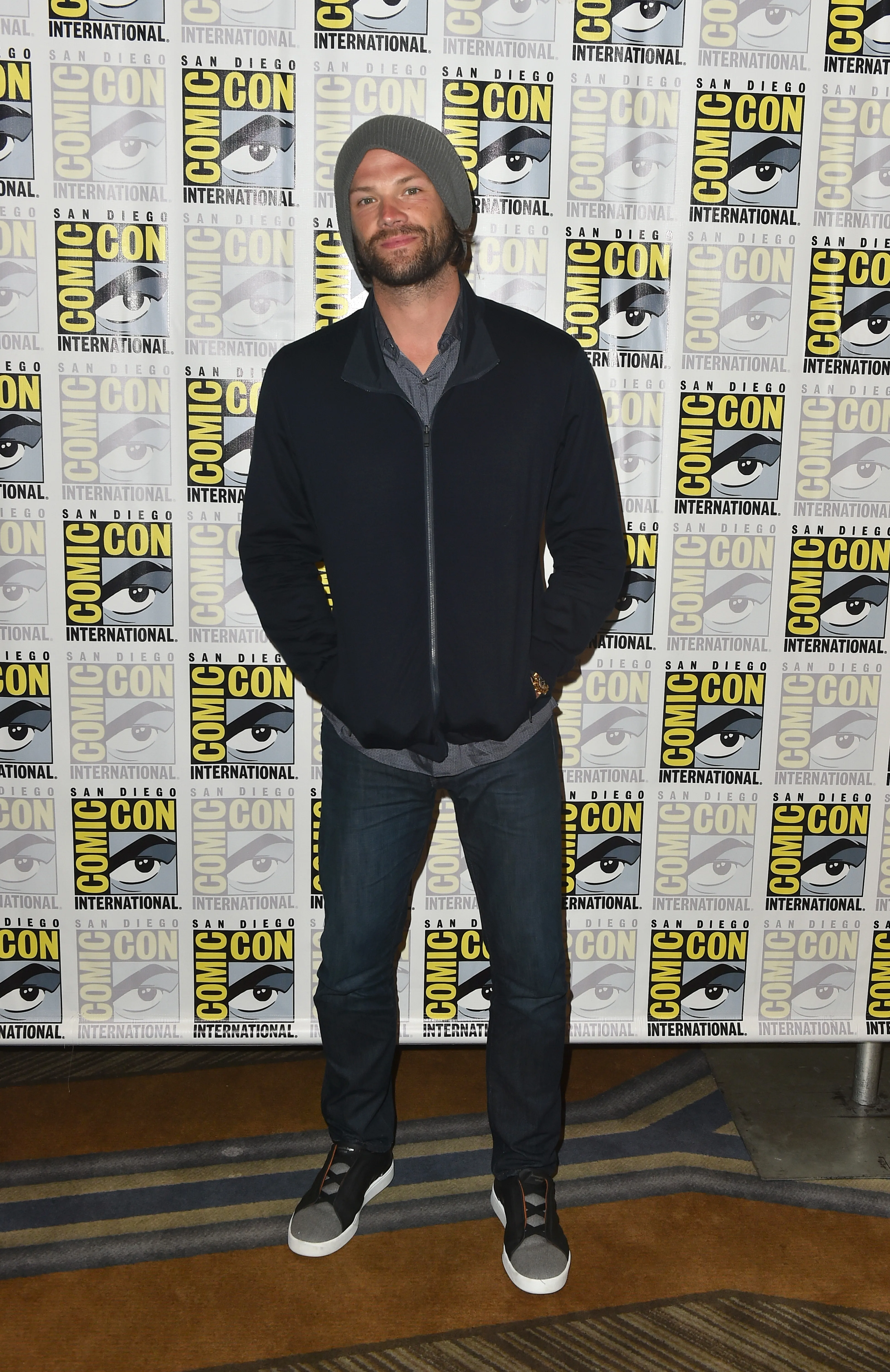 Jared Padalecki in a gray beanie, black zip-up jacket, jeans, and sneakers, stands smiling in front of a Comic-Con backdrop.