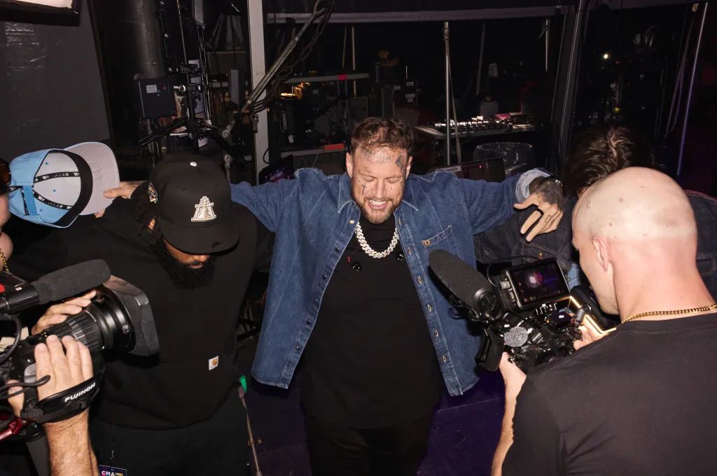 Jelly Roll with a wide grin and outstretched arms, surrounded by cameramen, backstage at CMA Fest.